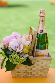 a bottle of champagne and flowers sit in a basket on a table with green grass