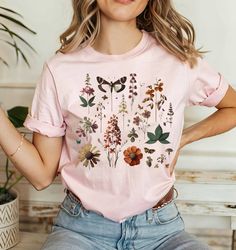 Discover our "Wildflower Graphic Tee" - a must-have Flower T Shirt that's as stylish as it is comfortable. Perfect for any plant lover or nature enthusiast, this tee makes for an exceptional gift for women who adore the cottagecore aesthetic or simply love to add a botanical touch to their wardrobe. Each shirt boasts a unique flower print, inspired by nature's wild beauty, making it the ideal graphic tshirt for casual wear or as a statement piece for the devoted plant lady. It's not just a t-shi Pink Graphic Tee With Plant Print, Pink Graphic Tee With Plants Print, Pink Crew Neck Top With Plant Print, Pink Cotton T-shirt With Plant Print, Graphic Tee With Plant Print Crew Neck, Pink T-shirt With Plants Print In Relaxed Fit, Trendy Crew Neck Tops With Plants Print, Spring Pink T-shirt With Plant Print, Botanical Style Screen Print Tops For Spring