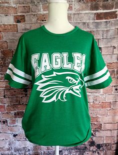 Introducing my Eagles school spirit lightweight jersey LAT tees, the perfect blend of softness, stretchiness, comfort, and cuteness! These tees are designed to elevate your school spirit wear to new levels of style and comfort. Crafted from high-quality lightweight material, these tees offer an exceptional softness that feels gentle against your skin. Embrace the comfort of these shirts, as they provide a cozy and luxurious feel that you'll never want to take off. The stretchy fabric ensures a perfect fit and allows for ease of movement. Whether you're cheering on your team or going about your day, these tees provide unrestricted comfort and flexibility. No more feeling constrained or uncomfortable in your school spirit wear! Shirts are unisex sizing - please see the size chart shown in th Collegiate Style School T-shirt With Screen Print, Green Sporty Cotton Shirt, School Spirit Tops For Football Season, Collegiate Letter Print T-shirt For School, School Spirit T-shirt For Game Day, College Football Season Short Sleeve T-shirt, Casual T-shirt For School Football Season, College T-shirt With School Spirit, Varsity Jersey T-shirt For Game Day