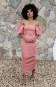 a woman standing in front of a stone wall with her hands on her hips wearing a pink dress