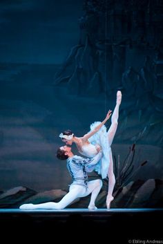 two ballerinas in white tutus and blue leotards, one holding the other's leg