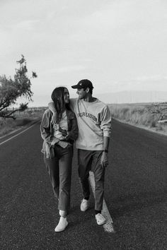 a man and woman walking down the middle of an empty road in black and white