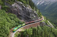 a train traveling down tracks next to a lush green forest filled mountain side covered in snow