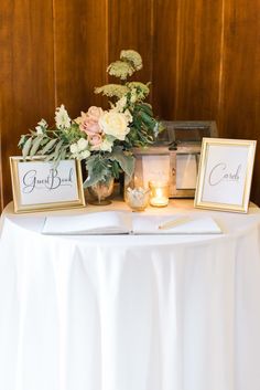 a table topped with flowers and two framed pictures next to a lit candle on top of a white cloth covered table