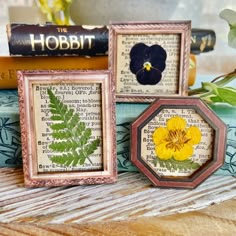 three framed pictures with flowers on them sitting on a table next to books and plants