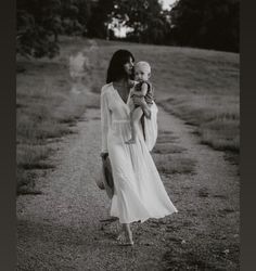 a woman walking down a dirt road holding a baby in her arms while wearing a white dress