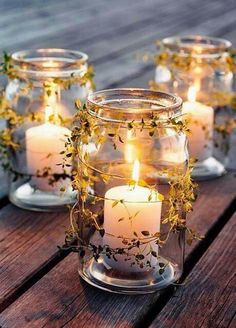 four glass jars filled with lit candles on top of a wooden table covered in greenery