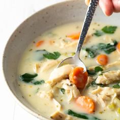 a person holding a spoon over a bowl of soup with chicken and carrots in it