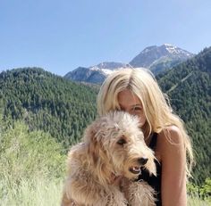 a blonde woman holding a white dog in front of a mountain range with trees and grass