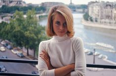 a woman with her arms crossed standing in front of a balcony overlooking a river and buildings