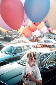 a man holding balloons in front of his face while standing next to a bunch of parked cars