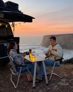 a man and woman sitting at a table in front of an suv with the ocean behind them