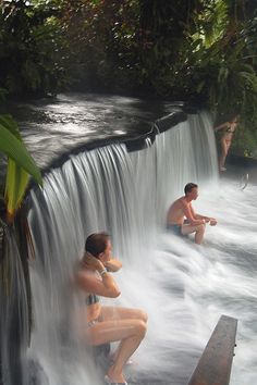 two people sitting in the middle of a waterfall while others stand around and bathe