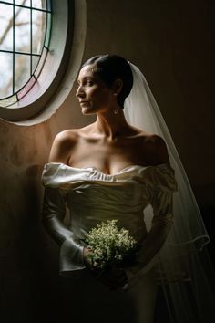 a woman standing in front of a window wearing a wedding dress and holding a bouquet