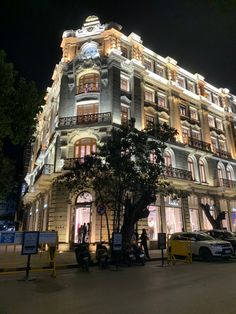 a large building lit up at night with cars parked in front of it and people standing on the sidewalk