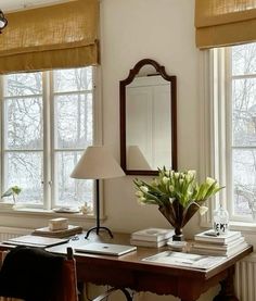 a desk with a mirror, lamp and flowers on it in front of two windows