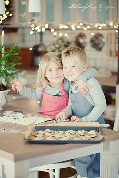 two children sitting at a kitchen table with food on the counter and christmas lights in the background