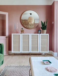 a living room with pink walls and white furniture in the corner, along with a large round mirror on the wall