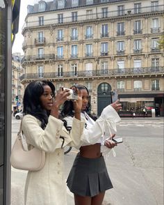 two women taking pictures with their cell phones in front of a building on the street