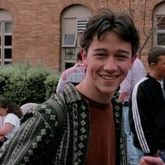 a young man standing in front of a brick building smiling at the camera with other people behind him