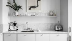 a white kitchen with marble counter tops and shelves filled with glasses, coffee maker, teapots and other items