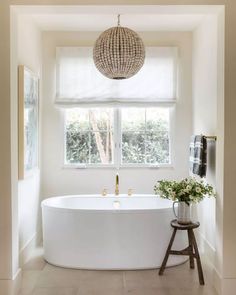 a white bath tub sitting under a window next to a wooden stool and potted plant