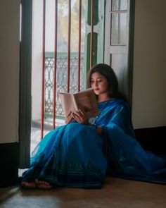 a woman sitting on the floor reading a book while wearing a blue sari and sandals