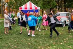 a group of people are dancing around in the grass near an old car and tent