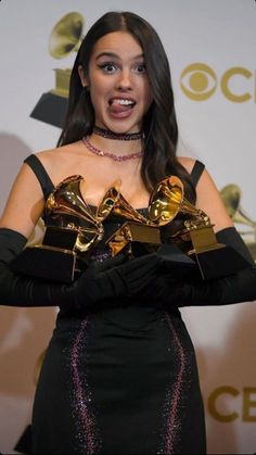 a woman in black dress holding two golden trophies and posing for the camera with her mouth open