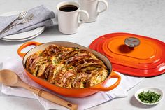 an orange casserole dish with meat and vegetables in it next to two cups of coffee