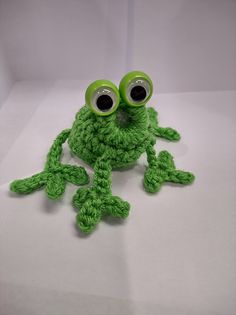 a green crocheted frog with big eyes sitting on top of a white surface