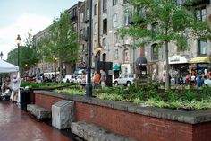 people are walking on the sidewalk in front of buildings and trees with plants growing out of them