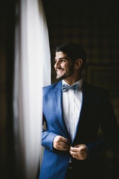 a man wearing a blue suit and bow tie standing next to a window with his hands in his pockets