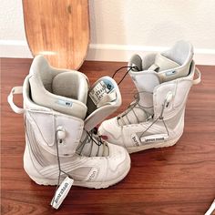 a pair of white snowboard boots sitting on top of a wooden floor next to a board