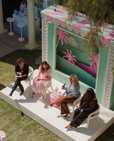 four women sitting on white chairs in front of a palm tree themed building with pink and green accents