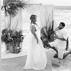 a man kneeling down next to a woman in a white dress on the beach with palm trees