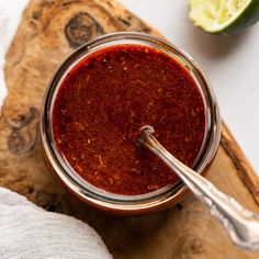 a jar filled with red sauce sitting on top of a wooden cutting board next to a lime