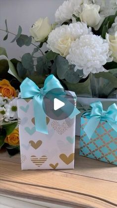 two gift bags sitting on top of a table with flowers in the background and a vase full of white carnations