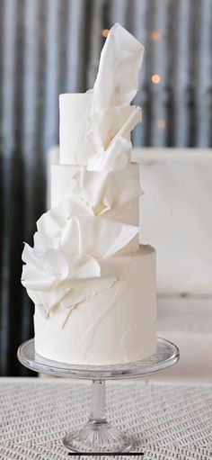 a white wedding cake with ruffles on the top is sitting on a glass stand