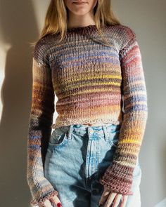 a woman standing in front of a wall with her hands on her hips wearing jeans and a colorful sweater