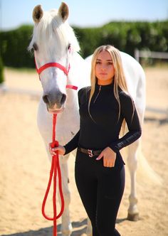 a woman standing next to a white horse on top of a dirt field with a red rope