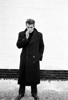 black and white photograph of a man standing in front of a wall wearing a trench coat