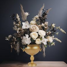 a gold vase with white flowers and feathers on a wooden table in front of a dark background