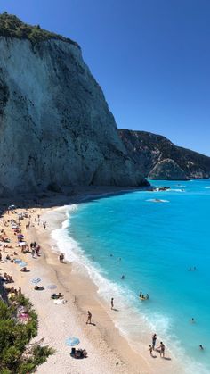 many people are on the beach and in the water near some cliffs with blue water