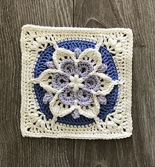 a crocheted square with a blue and white flower in the center on a wooden surface