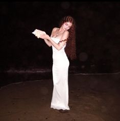 a woman in a white dress standing on the beach at night with her arms outstretched