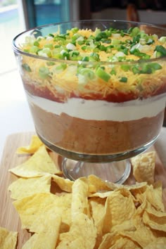 a glass dish filled with food on top of a wooden cutting board next to chips