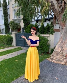 a woman in a yellow and blue dress standing next to a tree with her hand out