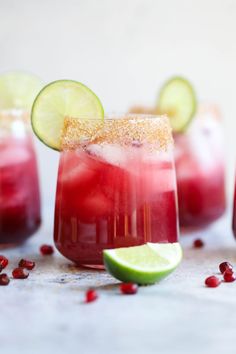 two glasses filled with red liquid and lime slices on the rims, surrounded by pomegranate