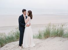 a man and woman standing on top of a sandy beach next to the ocean holding each other